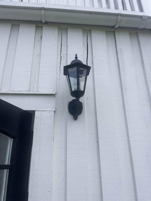 Vit fasad med spruckna panelplankor bredvid en svart lyktlampa under en balkong.