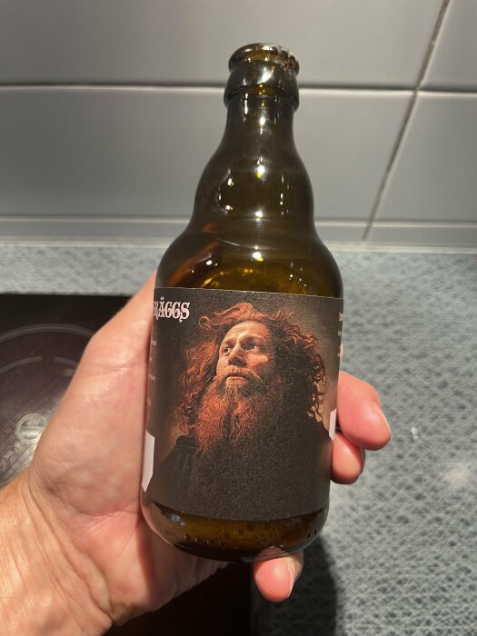 En hand som håller en flaska öl med etiketten Fader Rödskäggs XLIV-IPA Belgisk Ale, med en bild av en man med röd skägg och rött hår på etiketten.