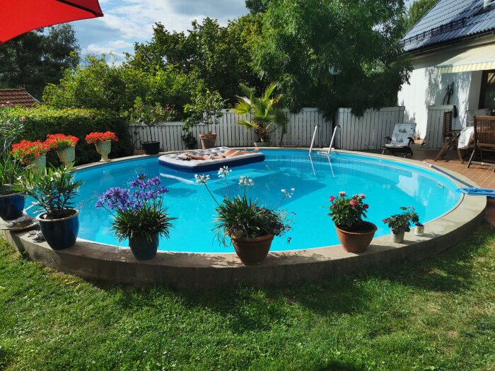 Rund pool i trädgård med blommor runt om och en person som solar på en madrass i vattnet. Sittplatser och växter på altan vid vit husfasad.