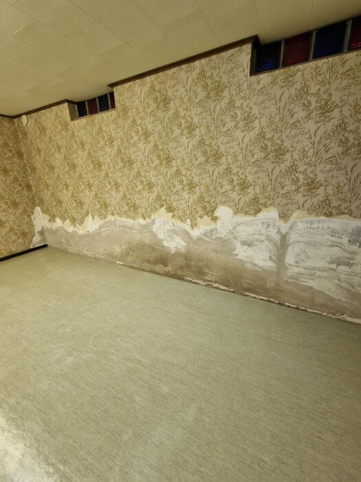 Wall in a basement with peeling and damaged wallpaper, partially covered with mold or water damage near the floor.