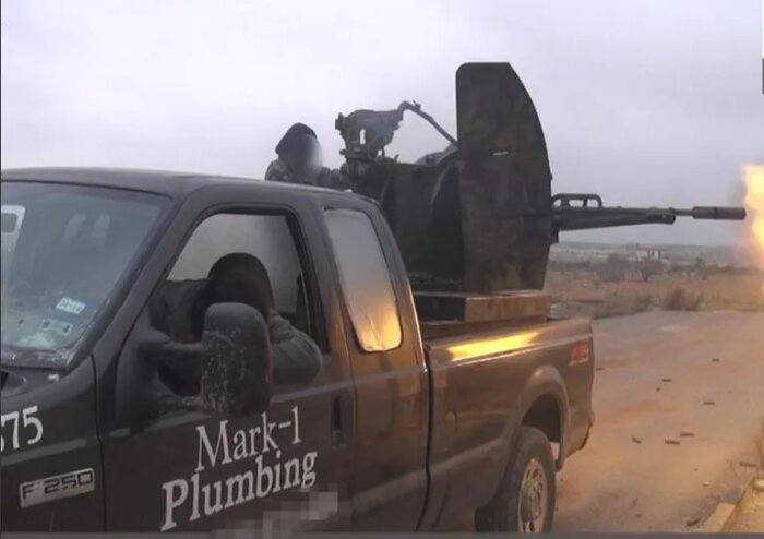 En svart pickup med texten "Mark-1 Plumbing" på sidan har ett tungt vapen monterat bak och en person som avfyrar det.