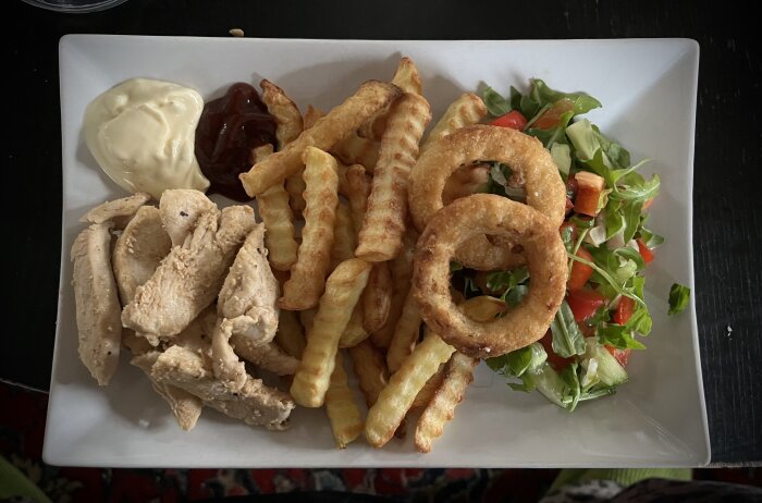 Tallrik med stekt kycklingfilé, krusig pommes frites, sallad med tomater, två lökringar, majonnäs och BBQ-sås.