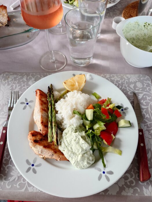 Tallrik med grillad kyckling, ris, kall basilikasås, sparris, och sallad samt ett glas rosévin och ett glas vatten på ett dukat bord.