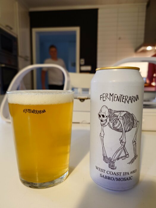 En ölburk med etiketten "Fermenterarna West Coast IPA #003 Sabro/Mosaic" och ett glas fyllt med öl, stående på ett bord.