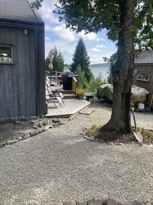 Grusgång vid ett hus med trädäck och utemöbler, omgiven av träd och sjö i bakgrunden. En trädgårdsspade står lutad mot ett träd.