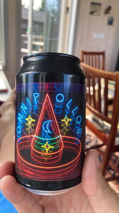 Svart aluminiumburk med färgglad, neoninspirerad design och texten "Omnipollo" hålls upp inomhus nära ett fönster.