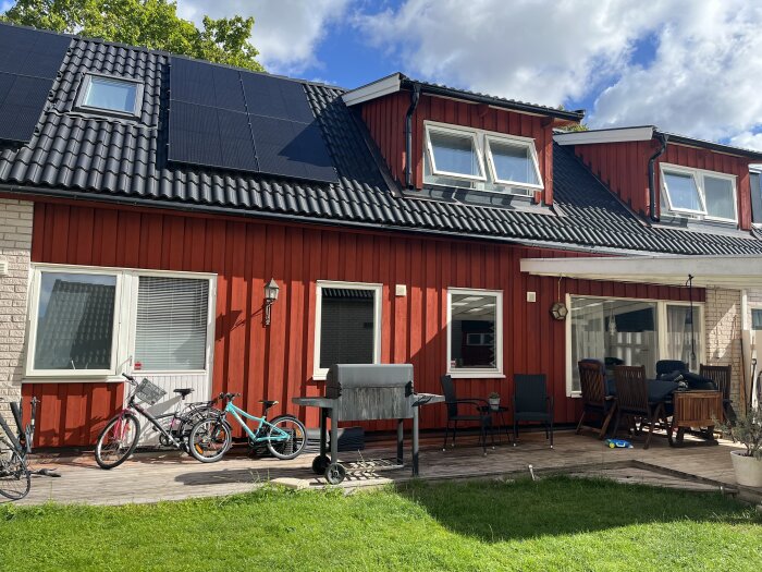 Rött hus med slamfärg och svarta takpannor, cyklar parkerade på en träaltan, utemöbler vid en täckt uteplats och solpaneler på taket.