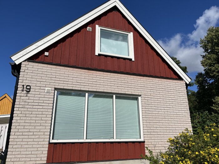 Ett hus med fasad delvis målad med röd slamfärg och delvis murverk. Fönster med persienner och gula blommor i trädgården.