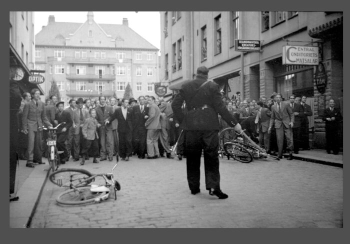 En svartvit bild av en folksamling på en gata framför en byggnad. En polis står vänd mot folkmassan, och omkullvälta cyklar ligger på marken.