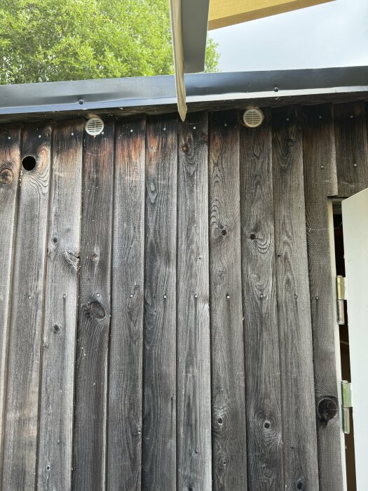 Sliten träfasad utomhus med två ventiler och en öppen dörr på höger sida.