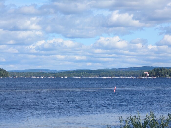 Sjön med småbåtar vid horisonten, skogklädda berg i bakgrunden, delvis molnig himmel och en röd boj i förgrunden.