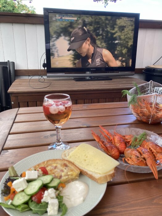 En tallrik med sallad, pajbit, smörgås och kräftor, samt ett glas dryck på ett träbord framför en TV som visar en golfspelare.