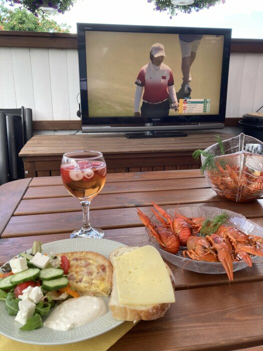 En TV som visar golf, en tallrik med sallad, paj och en smörgås med ost, ett glas med dryck och en skål kräftor på ett bord på en altan.