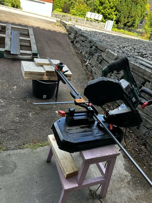 Bordssåg kapar metallstång i garageuppfart, omgiven av brädor och byggmaterial på pall. Grönska och stenmur i bakgrunden.