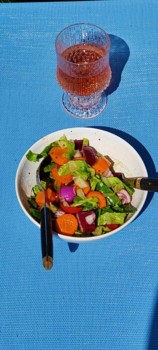 En sallad med färska grönsaker och en tallrik bredvid ett glas rosé på en blå duk.