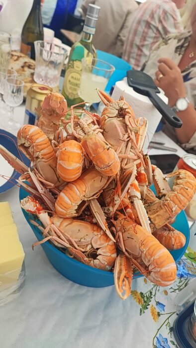 En blå skål fylld med kräftor på ett dukat bord, omgiven av dryckesglas, en vinflaska och en konservburk.