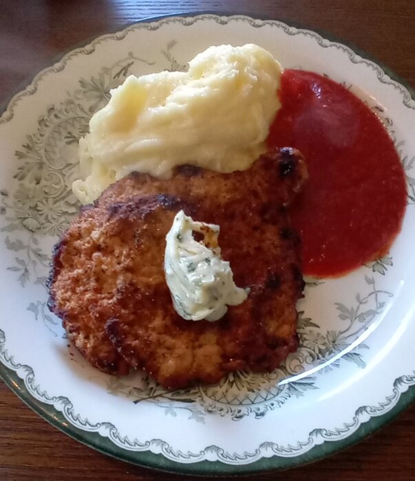 En tallrik med en schnitzel toppad med ört- och vitlökssmör, serverad med potatismos och en röd sås.