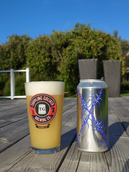 Ett glas med TIPA-öl från Omnipollo x Fidens bredvid en ölburk med blått taggtrådsmotiv på ett träbord utomhus, med buskar i bakgrunden.