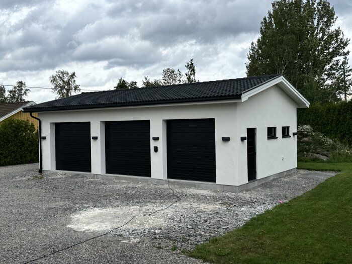 Vit byggnad med tre svarta garageportar omgiven av grus och en oavslutad infart framför.