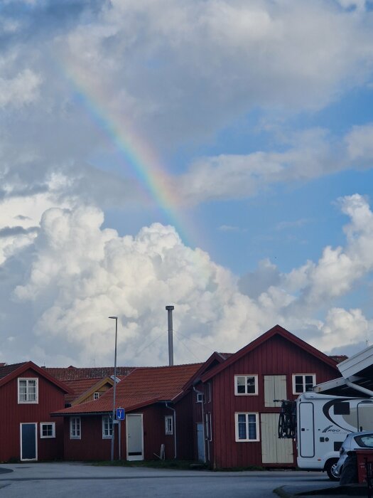 En regnbåge på en blå himmel med vita moln syns ovanför röda träbyggnader och en parkerad husbil i förgrunden.