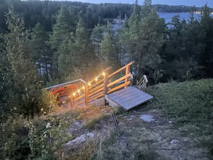 Belyst trätrappa med räcke och slingade 12v lampor, belägen i skogsmiljö med utsikt över en sjö vid skymning.