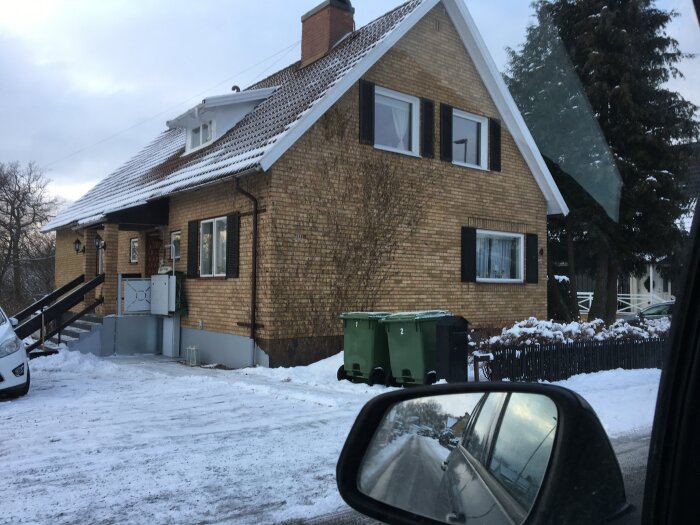 Gult tegelhus med snötäckt tak, grön soptunna, en vit bil parkerad på uppfarten och snö på marken, sett från bilens sido- och backspegel.