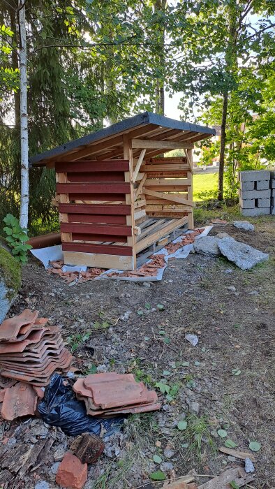 Nytt vedskjul med gles panel under byggnation, omgiven av träd och byggmaterial, redo att fyllas med ved.