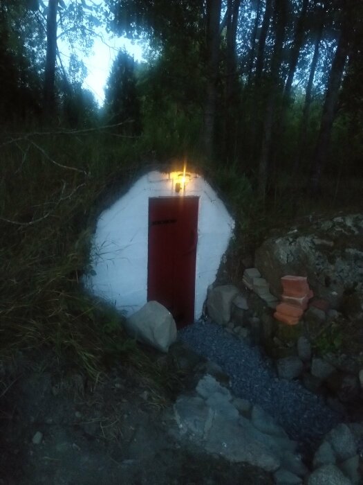 Vitputsad jordkällaringång med röd dörr och lyktbelysning i skymningen, omgiven av skog och stenar.