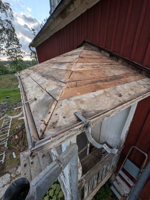 Entrétak med kuverttakskonstruktion och rödmålad vägg, förberett för att lägga ytpapp, synliga rännkrokar och trasig fotplåt. Stege och virkesrester i förgrunden.