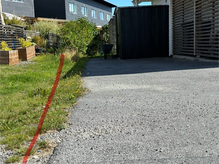 Gräsmatta med röd linje som markerar tomtgräns, intill asfalt och en carport. Träodlingslådor och buskar syns till vänster bortom tomtgränsen.