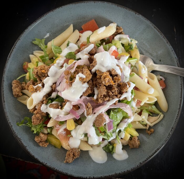 En tallrik med en pastasallad som innehåller penne pasta, blandad sallad, kyckling, grönsaker och tacorester, toppad med vit dressing.