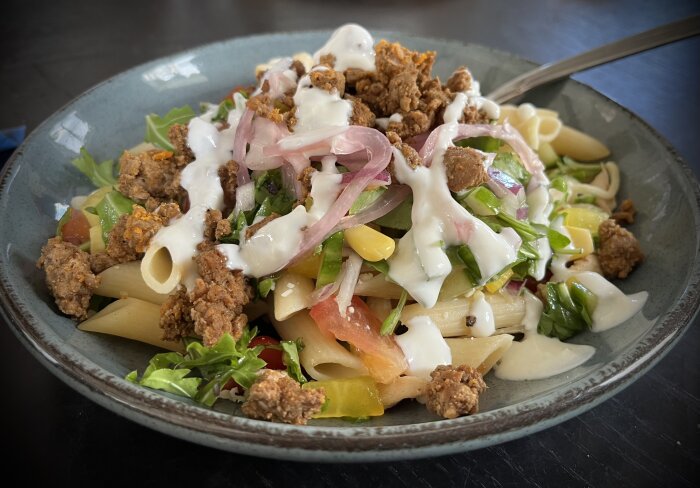 Pastasallad med kyckling, sallad, tomat, tacokött och dressing på ett grått keramikfat.