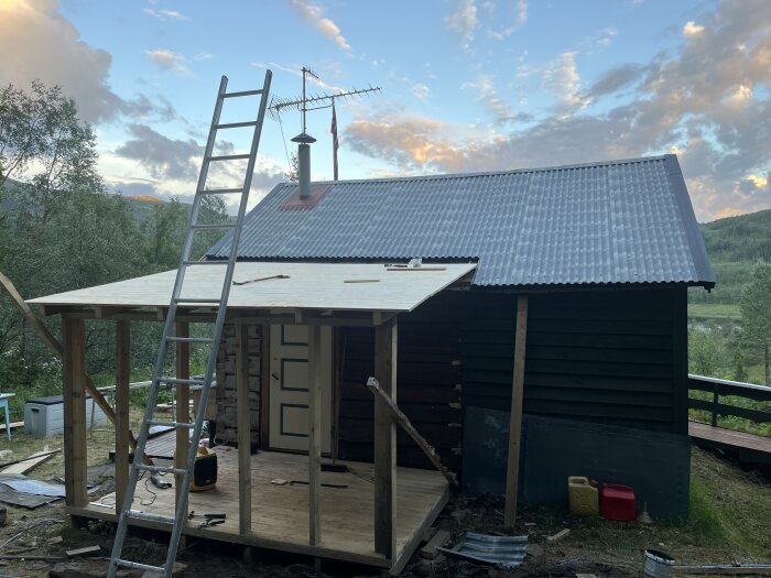 Fritidshus under renovering med obruten veranda och stege lutad mot taket. Bakgrunden visar skog och en molnig himmel. Byggmaterial spridda runt huset.