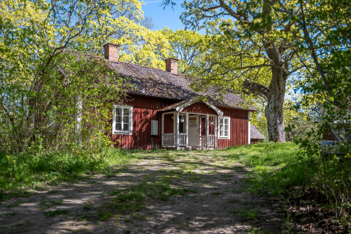 En gammal röd skolmästarbostad från 1700-talet med vita knutar omgiven av grönskande träd och växtlighet i vårens härliga solsken.