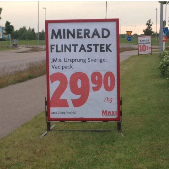 Affisch på gräsmatta vid trottoar som annonserar "Minerad flintastek, ursprung Sverige, 29,90 kr/kg" från Maxi, max 2 köp per hushåll.