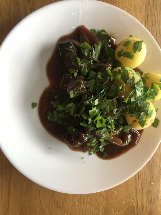 Boeuf bourguignon inspirerad gryta med kött, kantareller och små lökar, serverad med potatis och toppad med färsk persilja på en vit tallrik.