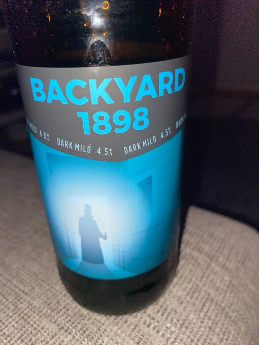 En flaska Backyard 1898 mörk öl med 4,5% alkoholhalt, etikettens design visar en silhuett av en person med hög hatt mot en blå bakgrund.