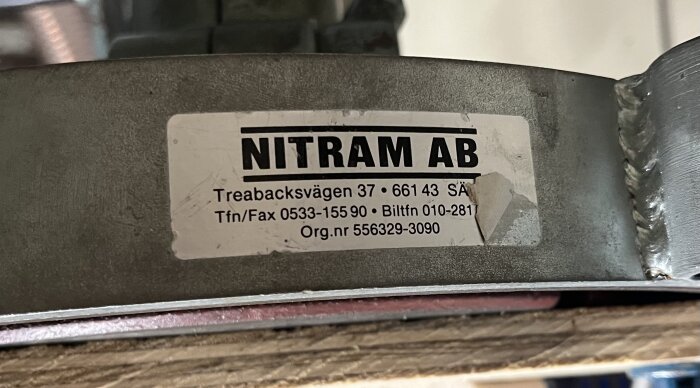 Label med kontaktinformation för Nitram AB, belägen på en metallkomponent. Adress: Treabacksvägen 37, 661 43 Sä. Telefonnummer: 0533-155 90 och 010-281.