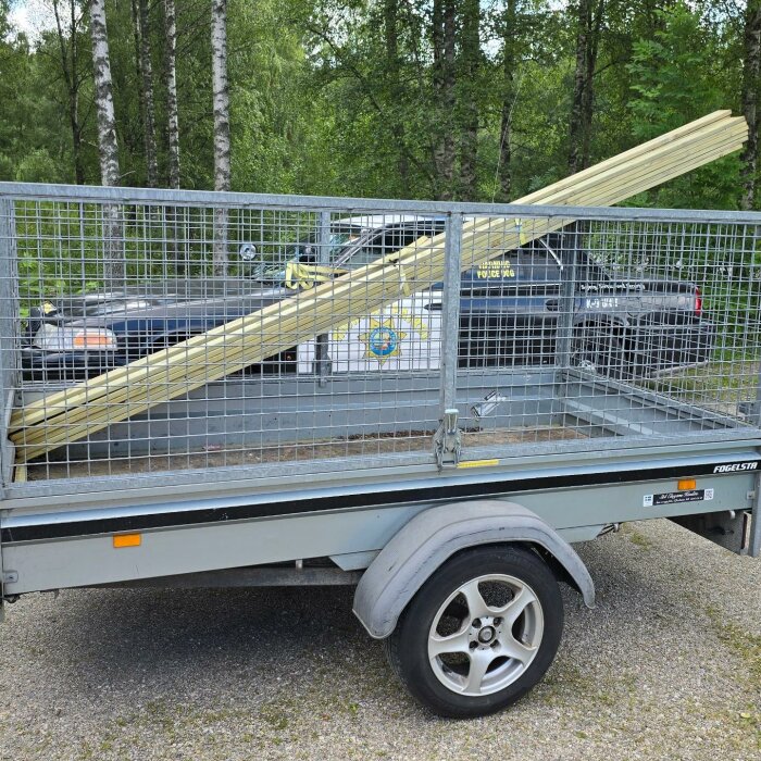 En släpkärra med nätväggar lastad med långa trälister, stående parkerad på grusväg bredvid två bilar, omgiven av skog.