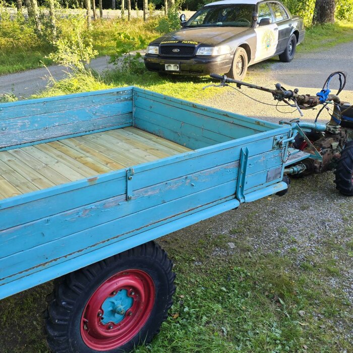 Blå träkärra med nytt trägolv kopplad till en tvåhjulstraktor står utomhus, med en polisbil parkerad i bakgrunden på en grusväg.