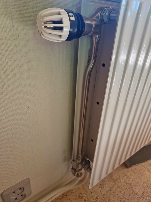 Närbild av en radiatorventil med en termostat, anslutna rör och en vit vägg i bakgrunden. Elementets lock är inte helt avskruvat.