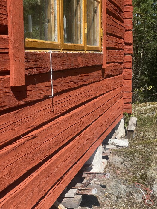 Timmerhus med röd fasad; gula träfönster saknar fönsterbläck och en bräda har placerats under fönstren, som visar tecken på fuktskador. Skadade timmerstockar syns.