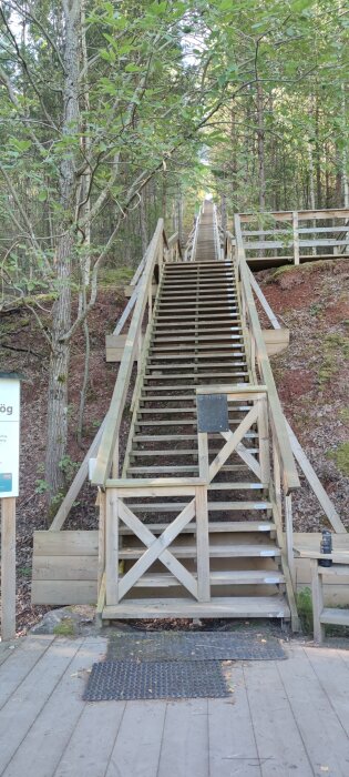 Trätrappa i skogsmiljö leder uppåt med ledstänger på båda sidor. Skylt till vänster om trappan och plankad mark vid trappans bas.