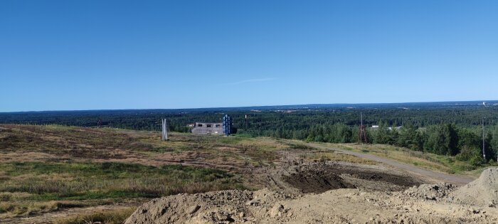 Vy över en pågående byggarbetsplats på ett fält med skog i bakgrunden under en klarblå himmel.