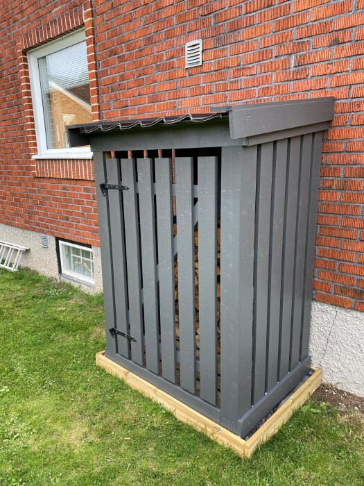 Vedbod med stående gråmålad spjälpanel och tak av korrugerad plåt, byggd vid tegelvägg med reglar av dimensionerna 45x70 och spjälor 22x120 eller 22x95.