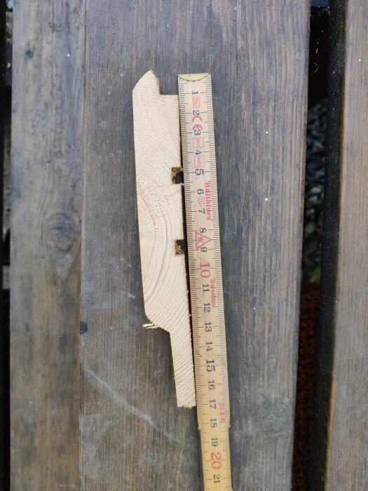 En bit träpanel med profilerad kant, ligger bredvid en trälinjal som mäter ungefär 20 cm, på en mörk träyta.