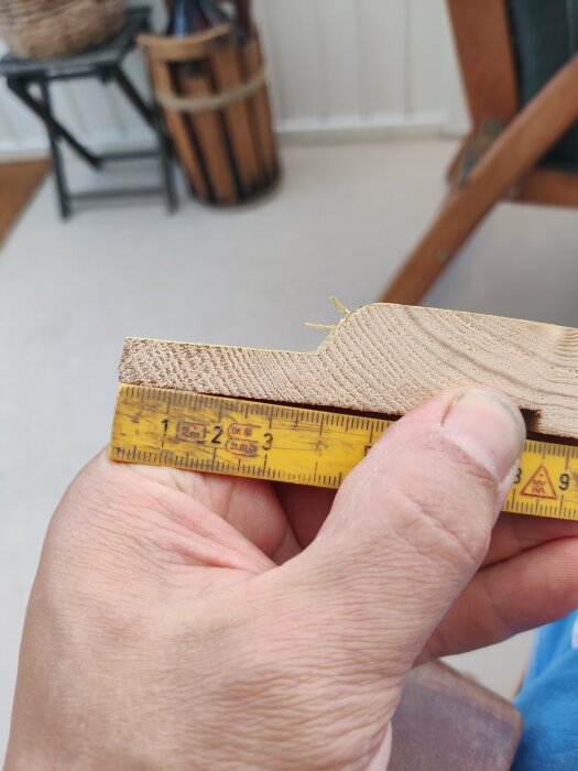 En hand håller en träbit och mäter dess tjocklek med en gul tumstock för att jämföra panelmått.