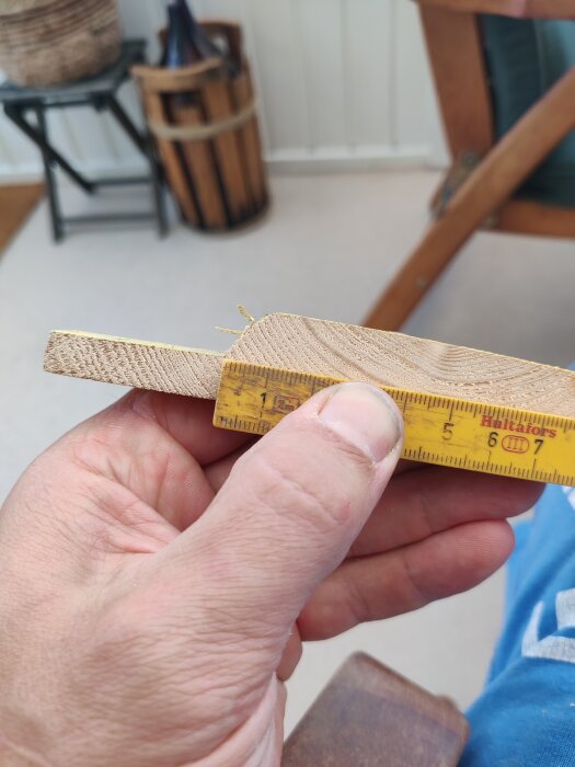 Närbild på en hand som håller en träbit mot en linjal för att mäta dess bredd, med en del av inomhusmiljö i bakgrunden.