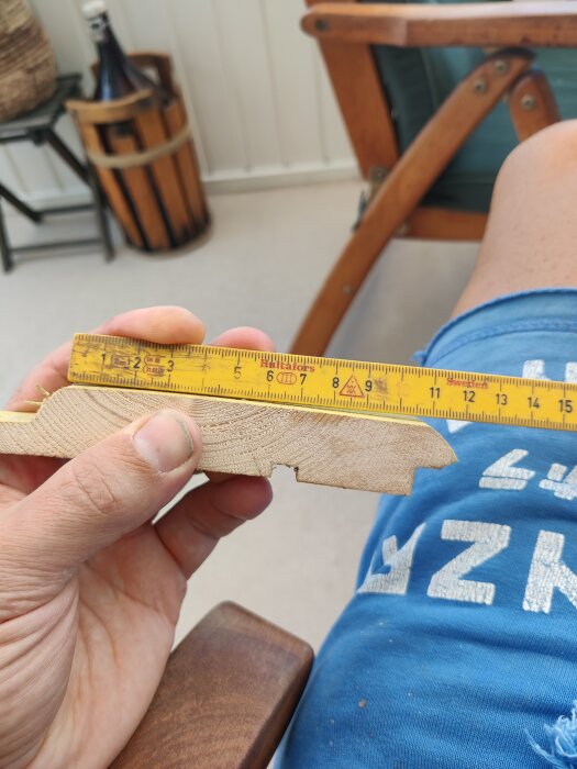 Närbild på en hand som håller en bit panel eller trälist med en måttstock på toppen för att mäta dess bredd, i bakgrunden syns ett ben och en trästol.