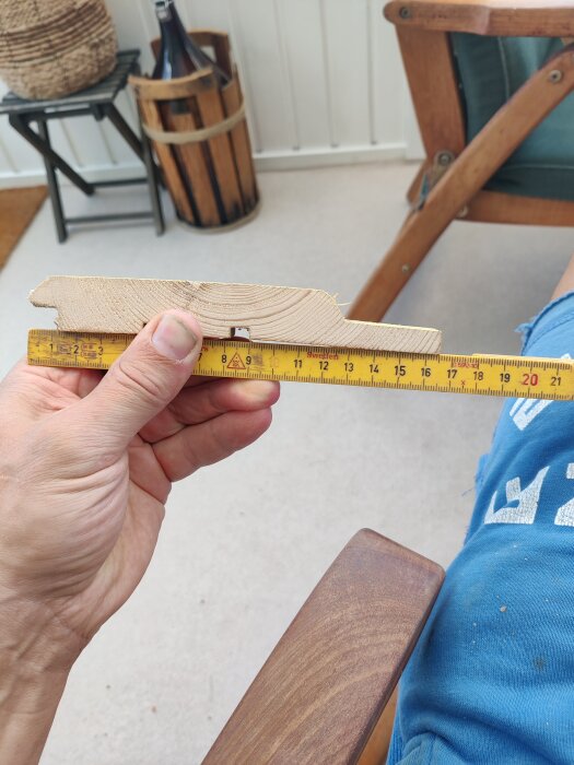 En hand håller och mäter en träpanel med en gul måttstock. Bakgrunden är ett rum med tve elevårig n cleasso köttberedning.